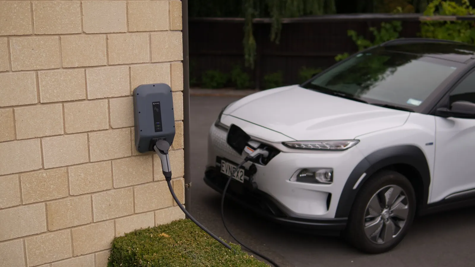 White electric SUV charging from a wall-mounted charger outside a house