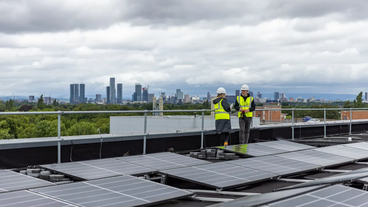 Two workers in safety vests on a rooftop solar array with a city skyline