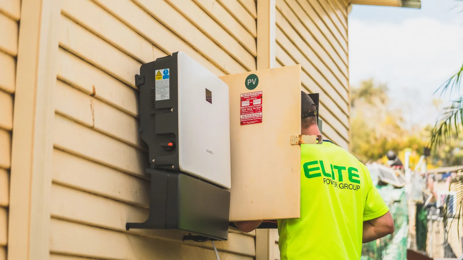 Home battery unit mounted on yellow house siding with an installer nearby
